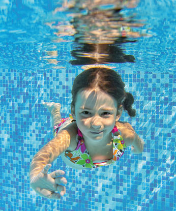 Ein Mädchen in buntem Badeanzug schwimmt unter Wasser in einem Pool mit blauen Mosaikfliesen und schaut lächelnd zur Kamera.