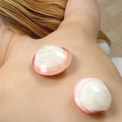 Eine Frau liegt mit dem Rücken nach oben auf einer Wellnessliege. Auf ihrem Rücken befinden sich zwei große, glänzende Muschelschalen. Die Muscheln sind rosa-weiß gemustert und wirken wohltuend auf der Haut.