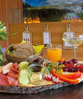Ein liebevoll angerichtetes Frühstücksbrett mit Aufschnitt, Käse, frischem Gemüse, Obst und Dips steht auf einem rustikalen Holztablett. Daneben ein Glas Orangensaft, ein Glas Sekt und ein Korb mit Körnerbrot. Im Hintergrund ein stilvoll eingerichteter Raum mit Holzverkleidung und dekorativen Schneidebrettern an der Wand.